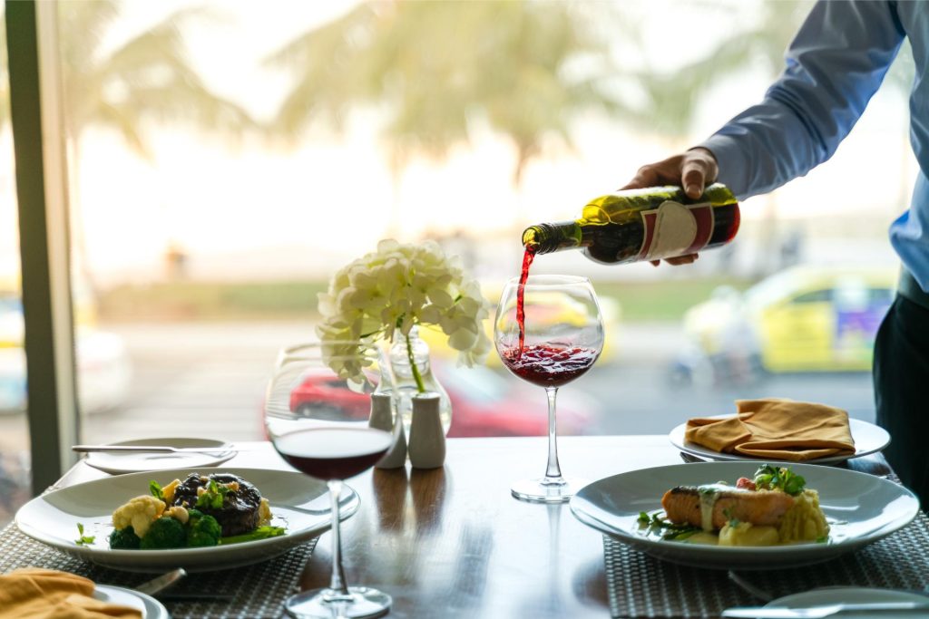 A waiter pours red wine into a glass at a restaurant table. There are two glasses of red wine and plates of food on the table. The restaurant has a large window with a view of a city street.