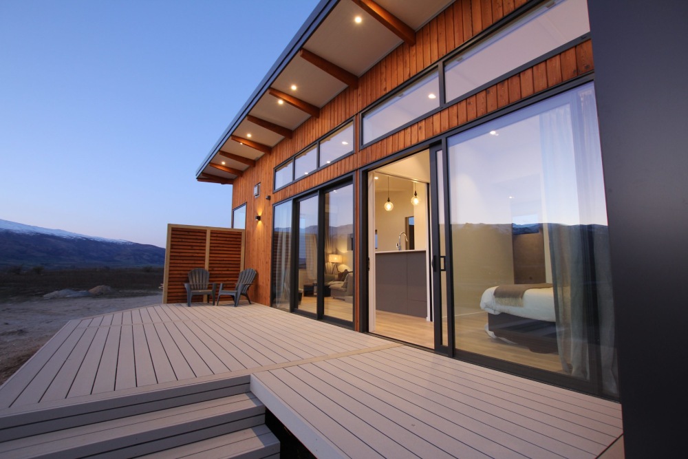 A modern, single-story cabin with a large wooden deck and sliding glass doors. The cabin has a dark exterior with wood paneling and large windows. A set of stairs leads down to the deck, and there are two chairs on the deck. Snow-capped mountains can be seen in the background.