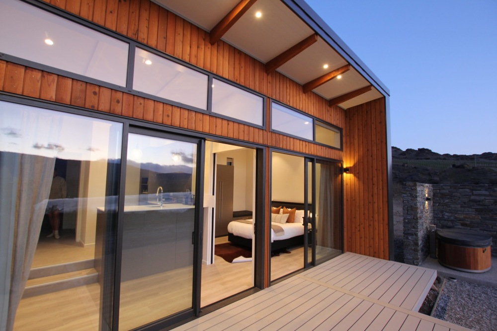 A modern, single-story cabin with a large wooden deck and sliding glass doors. The cabin has a dark exterior with wood paneling and large windows. A set of stairs leads down to the deck, and there are two chairs on the deck. A hot tub can be seen on the lower level, and the interior of the cabin is partially visible through the glass doors. Snow-capped mountains can be seen in the background.